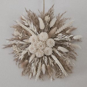 Könnte beinhalten: Ein kreisförmiges Trockenblumenarrangement mit einem zentralen Cluster cremefarbener Blüten, umgeben von beigen und braunen getrockneten Gräsern und Weizen. Aufgehängt an natürlichem Bindfaden. Heimdekoration.