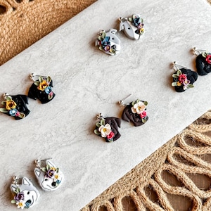 May include: Handmade polymer clay earrings featuring dog faces adorned with colorful floral arrangements. The earrings are in various colors, including black, brown, and white, with silver-colored earring hooks. The earrings are displayed on a light gray surface.