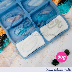Op de afbeelding: Een blauwe siliconen mal met zes ovale holtes, elk met een gedeeltelijk uitgeharde, lichtgekleurde hars. De mal is gemarkeerd met "Denver Silicone Molds" en bevat een roze cirkel met de aanduiding 80g. De holtes hebben het woord "JOKWINS" in reliëf.