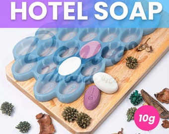 May include: Blue silicone mold with 18 individual cavities for making hotel soap bars. The mold is on a wooden surface with three small soap bars, one white, one purple, and one pink. The text "10g" is visible in the lower right corner of the image.