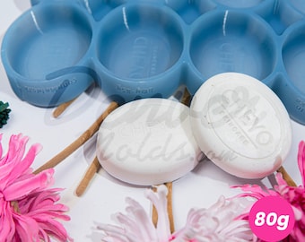 May include: A light blue silicone mold with circular cavities, next to two white oval soaps. The soaps have the text "CHEYO SKINCARE" and are labeled with 80g. Pink and white flowers are in the foreground.