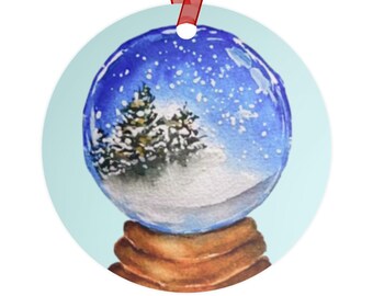 Op de afbeelding: Ronde kerstversiering met een aquarel sneeuwbolontwerp. De bol heeft een blauwe lucht met sneeuwval, een besneeuwd landschap en dennenbomen. De versiering is bevestigd aan een rood lint.