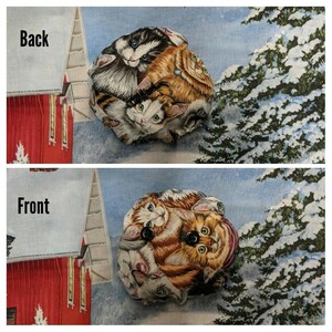 May include: A round fabric ornament featuring a design of several cats in different poses. The cats are brown, white and black with green and blue eyes. The ornament is displayed against a snowy backdrop with a red barn in the background.