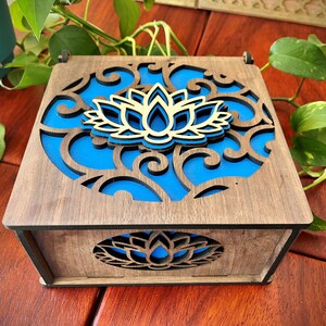 May include: A wooden box with a blue and gold lotus flower design. The box has a hinged lid and a cutout design on the front.