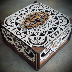 May include: A decorative wooden box with a detailed, layered design. The lid features a white, mandala-style pattern with a lotus flower accent. The box has a dark brown base and white accents, creating a contrast.