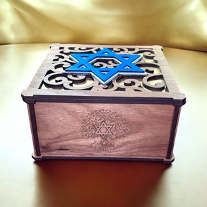 May include: A wooden box with a blue Star of David cutout on the top and a tree of life design engraved on the front. The box is made of dark wood and has a decorative, intricate design.