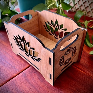 May include: A wooden storage box with a natural wood finish and black trim. The box has a cutout design of a flower on each side.