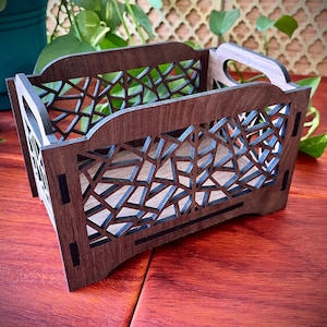 May include: A dark brown wooden box with a geometric pattern cut out of the sides. The box has a handle on the top and is sitting on a wooden surface.