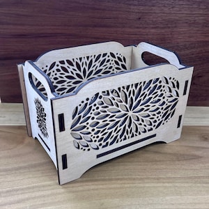 May include: A rectangular light-colored wooden storage crate featuring intricate laser-cut floral patterns on the sides and ends. The box has integrated handles on the two shorter sides and dark, burnt-edge detailing around the cutouts.