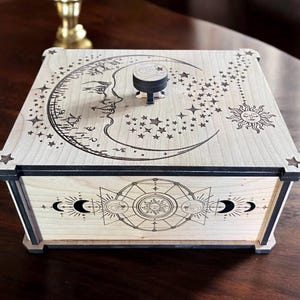 May include: A wooden box with a black trim and a decorative design of a crescent moon, stars, and a sun. The box has a small knob on the top.