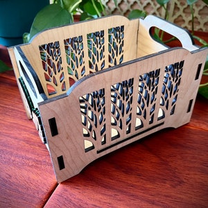 May include: A wooden storage box with a natural finish. The box features cut-out tree designs on the sides and a handle. The interior is visible through the cutouts. The box is rectangular and sits on a wooden surface.