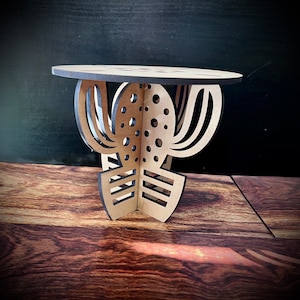 May include: A wooden table with a cactus design. The table is made of light brown wood and has a round top. The cactus design is cut out of the wood and is painted black.