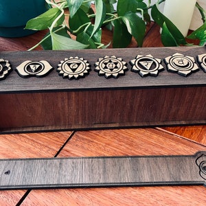 May include: Wooden incense holder with seven chakra symbols engraved on the top. The holder is brown and has a matching incense stick resting on the bottom.