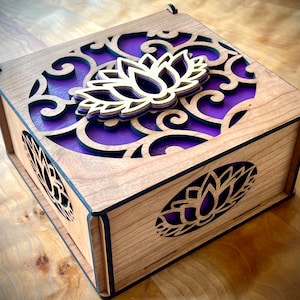 May include: A wooden box with a purple and gold lotus flower design. The box is made of cherry wood and has a black finish. The lid of the box is open and the inside is visible.