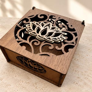 May include: A square wooden box featuring a dark brown finish with a laser-cut lid. The lid displays an intricate circular filigree pattern surrounding a central layered lotus flower design. A matching lotus cutout is visible on the front side panel.