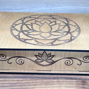 May include: A wooden box with a black trim and a laser-cut design of a lotus flower mandala on the lid. The box has a floral design on the front.