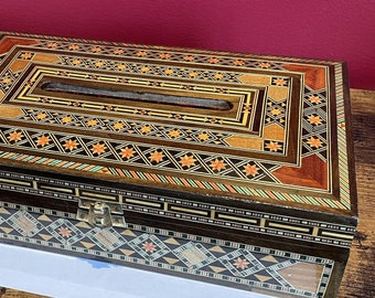 May include: A rectangular wooden box with intricate inlaid patterns in shades of brown, orange, and black. The box features a hinged lid and a rectangular opening on top. Decorative geometric patterns adorn the box's surface. The box is placed on a wooden table.