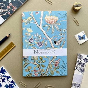 Op de afbeelding: Een blauw notitieboek met een bloemenpatroon en een wit label met de tekst "LAURA SOWERBY NOTEBOOK".