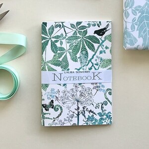 May include: A white notebook with a green and blue floral design. The cover features a small bird and the text "LAURA SOWERBY NOTEBOOK".