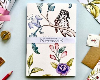 May include: A white notebook with a floral design and a bird on a branch. The notebook has the text "LAURA SOMERVILLE NOTEBOOK" printed on the front.
