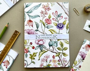 May include: A white notebook with a floral design featuring pink, purple, and green flowers. The notebook has a white label with the text "LAURA SOWERBY - NOTEBOOK".