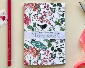 May include: A white notebook with a floral design in shades of pink, green, and black. The notebook has a black bird on the front and the text "LAURA SOWERBY NOTEBOOK" printed on a white band.