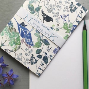 Peut inclure: Un carnet blanc avec un motif floral et un oiseau. La couverture présente un oiseau noir, des feuilles vertes et des fleurs bleues. Le texte "LAURA SOWERBY NOTEBOOK" est imprimé sur la couverture.
