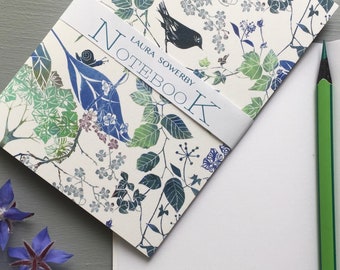 May include: A white notebook with a floral and bird design. The cover features a black bird, green leaves, and blue flowers. The text "LAURA SOWERBY NOTEBOOK" is printed on the cover.