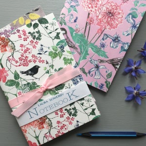 Può includere: Due quaderni con motivi floreali con copertine rosa e bianche. Il quaderno a sinistra ha un uccello nero e il testo "LAURA SOWERBY NOTEBOOK". Il quaderno a destra ha uno sfondo rosa con un motivo floreale. Una matita blu è su una superficie grigia.