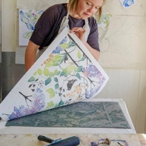 May include: A woman is using a printing press to create a floral design. She is holding a sheet of paper with a colorful floral pattern printed on it. The paper is being pressed onto a printing plate with a roller. There are tools and paint on the table.