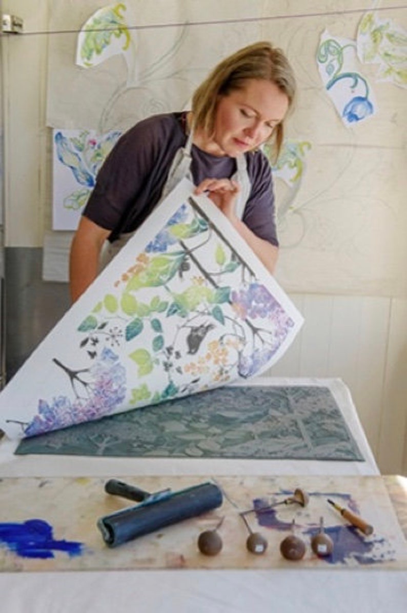 May include: A woman is using a printing press to create a floral design. She is holding a sheet of paper with a colorful floral pattern printed on it. The paper is being pressed onto a printing plate with a roller. There are tools and paint on the table.