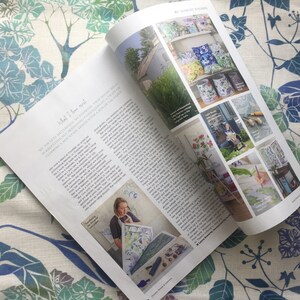 May include: A magazine spread featuring a woman painting a floral design on a large sheet of paper. The magazine article discusses the artist's love of nature and her creative process. The article also includes photos of the artist's work, including floral-themed wallpaper, cushions, and lampshades.