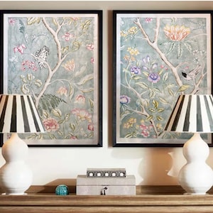 May include: Two framed prints with black frames and a floral design in shades of blue, green, pink, and yellow. The prints feature birds and branches. Two white lamps with black and white striped shades are on a wooden table in front of the prints.
