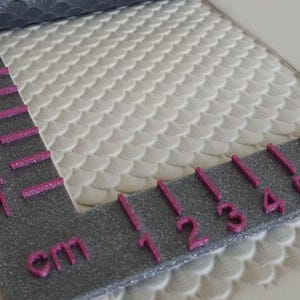 Könnte beinhalten: Nahaufnahme einer strukturierten, cremefarbenen Oberfläche mit einem schuppenartigen Muster. Ein graues Lineal mit pinkfarbenen Glitzer-Markierungen und Zahlen in Zentimetern ist am Rand positioniert. Das Lineal zeigt Messungen bis zu 4 cm an.