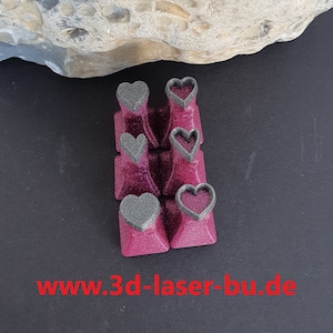 Könnte beinhalten: Satz von acht herzförmigen Keycaps in dunkelrosa mit schwarzem und silbernem Glitzerfinish. Die Keycaps haben einen herzförmigen Ausschnitt in der Mitte, der ein schwarzes Herz freilegt.