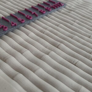Könnte beinhalten: Nahaufnahme eines strukturierten, hellbeigen Stoffes mit einem sich wiederholenden, wellenförmigen Muster. Ein dunkelgraues Lineal mit rosa Zahlen von 1 bis 9 liegt diagonal über dem Stoff und dient als visuelle Referenz für Maßstab und Messung. Die Stoffstruktur ähnelt Bambus.