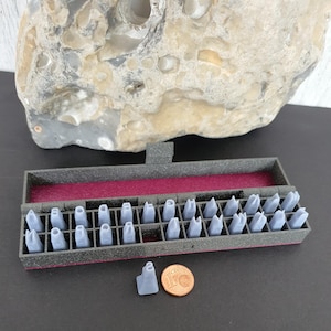 Könnte beinhalten: Ein Satz grauer Buchstabenstempel in einem schwarzen Kasten mit bordeauxrotem Innenraum. Ein einzelner Stempel und eine Euro-Münze befinden sich neben dem Kasten. Ein großer, grauer Felsen ist im Hintergrund.