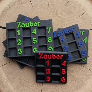 Könnte beinhalten: Eine Sammlung schwarzer quadratischer Puzzleteile mit dem Wort "Zauber" und Zahlen in Grün, Blau und Rot. Die Teile sind auf einer Holzoberfläche angeordnet und zeigen das Design des Spiels.