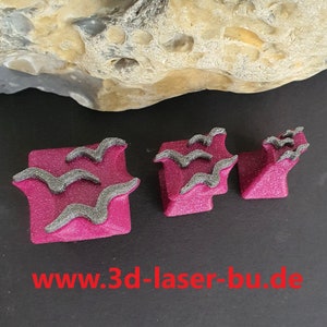 Könnte beinhalten: Drei rosa Stempel aus Gummi mit einem schwarzen und grauen Design von Vögeln, die in einer Schar fliegen. Die Stempel befinden sich auf einer schwarzen Oberfläche. Der Text "www.3d-laser-bu.de" ist am unteren Rand des Bildes sichtbar.