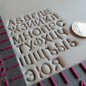 Könnte beinhalten: Nahaufnahme eines grauen Lederstempels mit kyrillischen Buchstaben. Die Buchstaben sind in das Leder geprägt. Ein Lineal und eine Euro-Münze sind ebenfalls sichtbar. Der Stempel wird für Lederarbeiten und die Gestaltung individueller Designs verwendet.