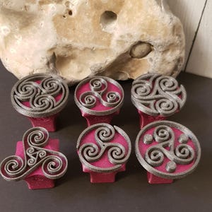 Könnte beinhalten: Sechs dekorative Schrankknöpfe mit einem keltischen Spiraldesign. Die Knöpfe haben ein metallisch graues Spiralmuster auf einer kreisförmigen, magentafarbenen Basis. Die Knöpfe sind in einer Gruppe angeordnet, mit einem Naturstein im Hintergrund.