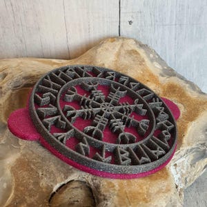 Könnte beinhalten: Runder, dunkelgrauer Ausstecher mit einem Schneeflockenmuster und Runensymbolen. Der Ausstecher hat eine dunkelrosa Basis und einen Griff. Das detaillierte Design eignet sich für thematische Kekse oder Gebäck. Durchmesser: ca. 10 cm.