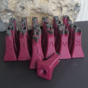 Könnte beinhalten: Sammlung burgunderfarbener, burgförmiger Stempel mit grauen Oberteilen. Die Stempel sind in Reihen angeordnet, einige zeigen geätzte Symbole. Ein Stempel ist abgelöst und zeigt ein Loch. Der Hintergrund ist ein hellfarbiger, strukturierter Stein.