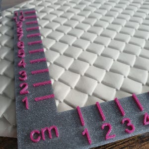 Könnte beinhalten: Nahaufnahme eines weißen, strukturierten Materials mit einem gewebten Muster. Ein graues Lineal mit rosa Markierungen und Zahlen in Zentimetern liegt auf dem Material und misst bis zu 5 cm. Das Lineal hat auch die Buchstaben "cm".