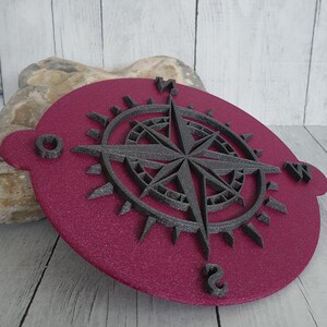 Könnte beinhalten: Ein weinrotes Kompassrosen-Design mit strukturiertem Finish. Die Kompassrose ist dunkelgrau und weist einen detaillierten Stern und Richtungsmarkierungen auf. Das Design ist auf einer weinroten, ovalen Basis mit strukturierter Oberfläche angebracht.
