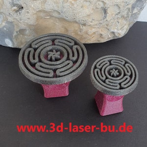 Könnte beinhalten: Zwei graue und schwarze 3D-gedruckte runde Stempel mit einem Labyrinth-Design und einem Stern in der Mitte. Die Stempel sind auf roten Sockeln montiert.