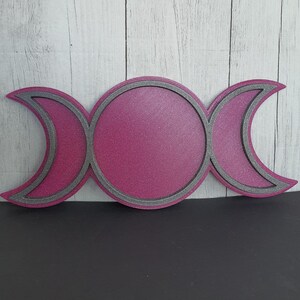 Könnte beinhalten: Ein magenta-farbenes Triple-Moon-Symbol mit strukturierter Oberfläche. Das Design zeigt einen zentralen Kreis, der von zwei Halbmonden flankiert wird, alle grau umrandet. Das Symbol befindet sich vor einem hellen Holzmaserungshintergrund.