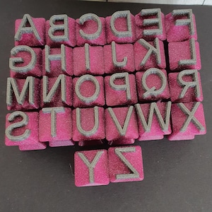 Könnte beinhalten: Ein Satz Alphabetstempel, jeder mit einem grauen Buchstaben auf einer pinkfarbenen, glitzernden quadratischen Basis. Die Buchstaben sind in einer serifenlosen Schriftart gehalten. Die Stempel sind in Reihen und Spalten angeordnet und zeigen das gesamte Alphabet.