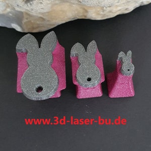 Könnte beinhalten: Drei graue, glitzernde Ausstecher in Hasenform. Die Ausstecher sind in verschiedenen Größen und auf einer rosa Oberfläche angeordnet. Der größte Ausstecher befindet sich links, der kleinste rechts und der mittlere Ausstecher in der Mitte. Die Website-Adresse www.3d-laser-bu.de ist unter den Ausstechern aufgedruckt.