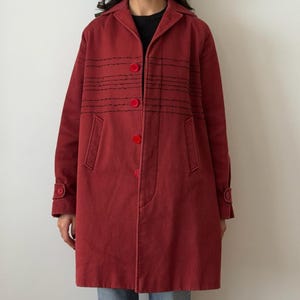 Può includere: Un cappotto rosso con cuciture nere e bottoni rossi. Il cappotto ha un colletto, maniche lunghe e tasche. Il cappotto, lungo circa 90 cm, sembra essere realizzato in un tessuto robusto.